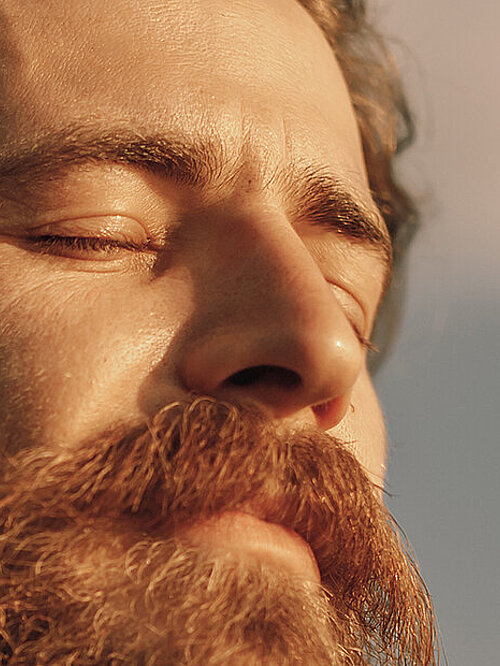 From below side view of tranquil peaceful adult bearded male in checkered shirt standing with eyes closed against cloudy sky in sunny day